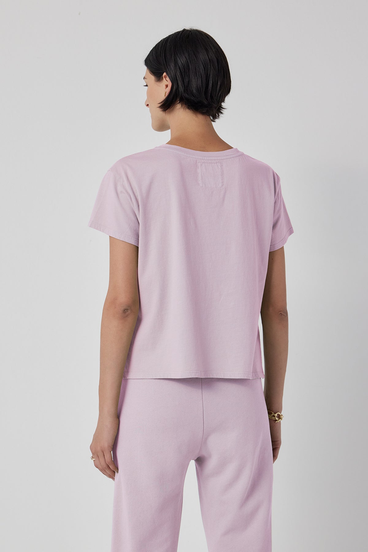   A person with short dark hair is shown from the back, wearing a Velvet by Jenny Graham TOPANGA TEE in light purple, styled in a relaxed fit with short sleeves, accompanied by matching pants. 