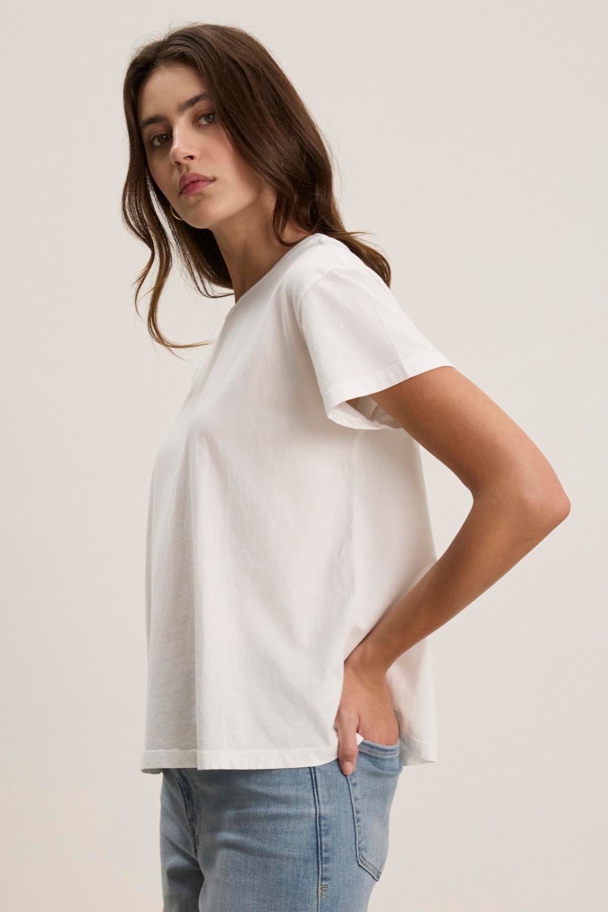   A person wearing the relaxed-fit, white TOPANGA TEE by Velvet by Jenny Graham, paired with blue jeans, stands sideways with hands in pockets, gazing forward. 