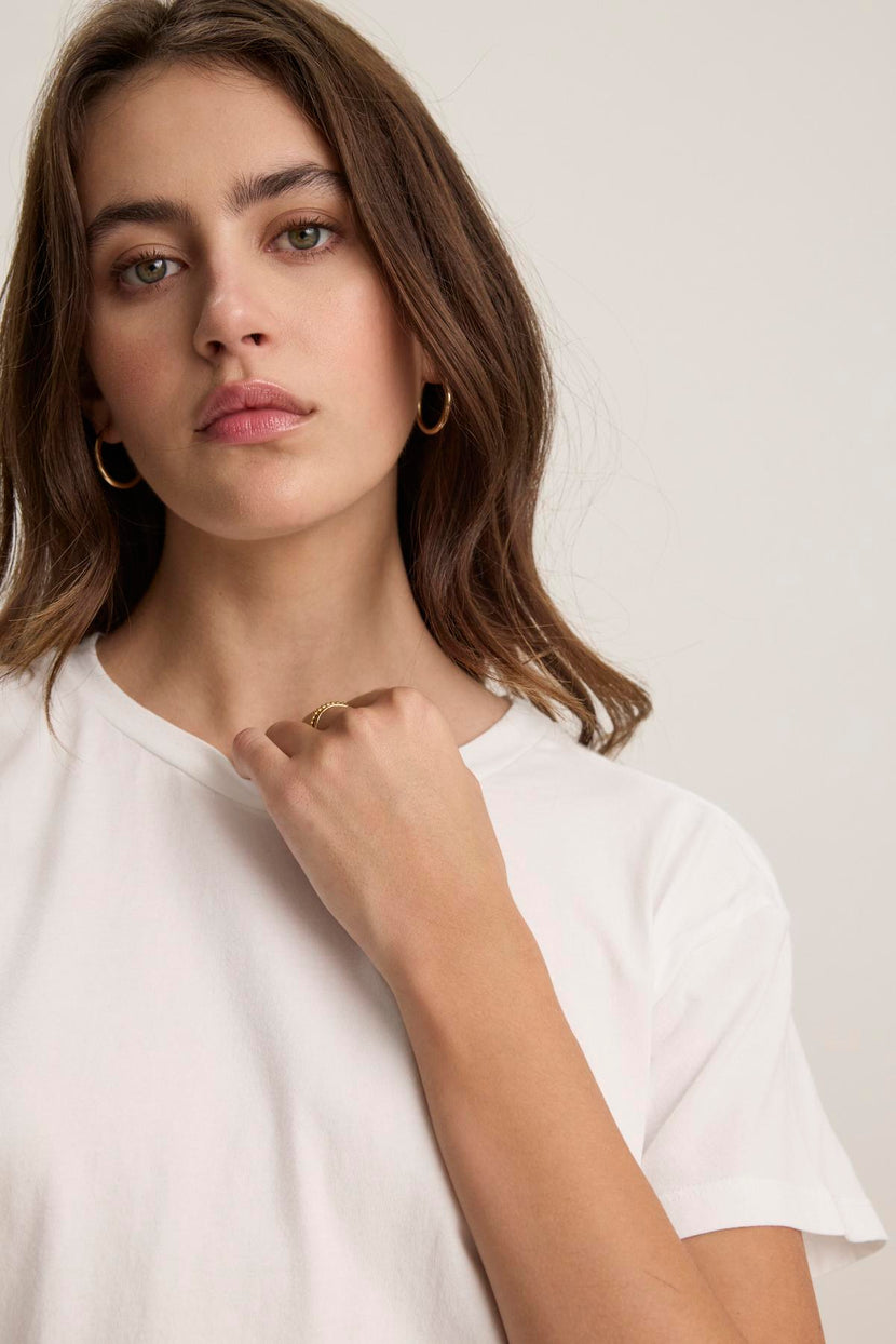 A person with long hair and hoop earrings wears the TOPANGA TEE, a relaxed-fit, organic cotton white t-shirt from Velvet by Jenny Graham. They are looking forward with a neutral expression and have their hand near their chin.