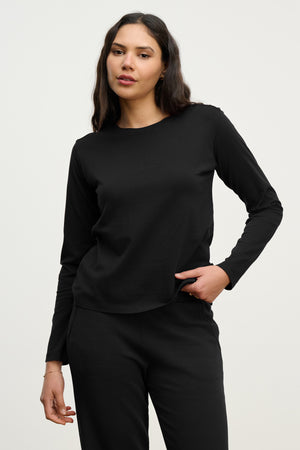 A woman with long dark hair wears the Velvet by Jenny Graham VICENTE TEE in black, paired with matching pants, standing against a plain light background.