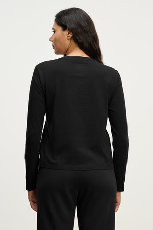A woman with long dark hair is seen from behind, wearing the Velvet by Jenny Graham VICENTE TEE in black with black pants, set against a plain light background.