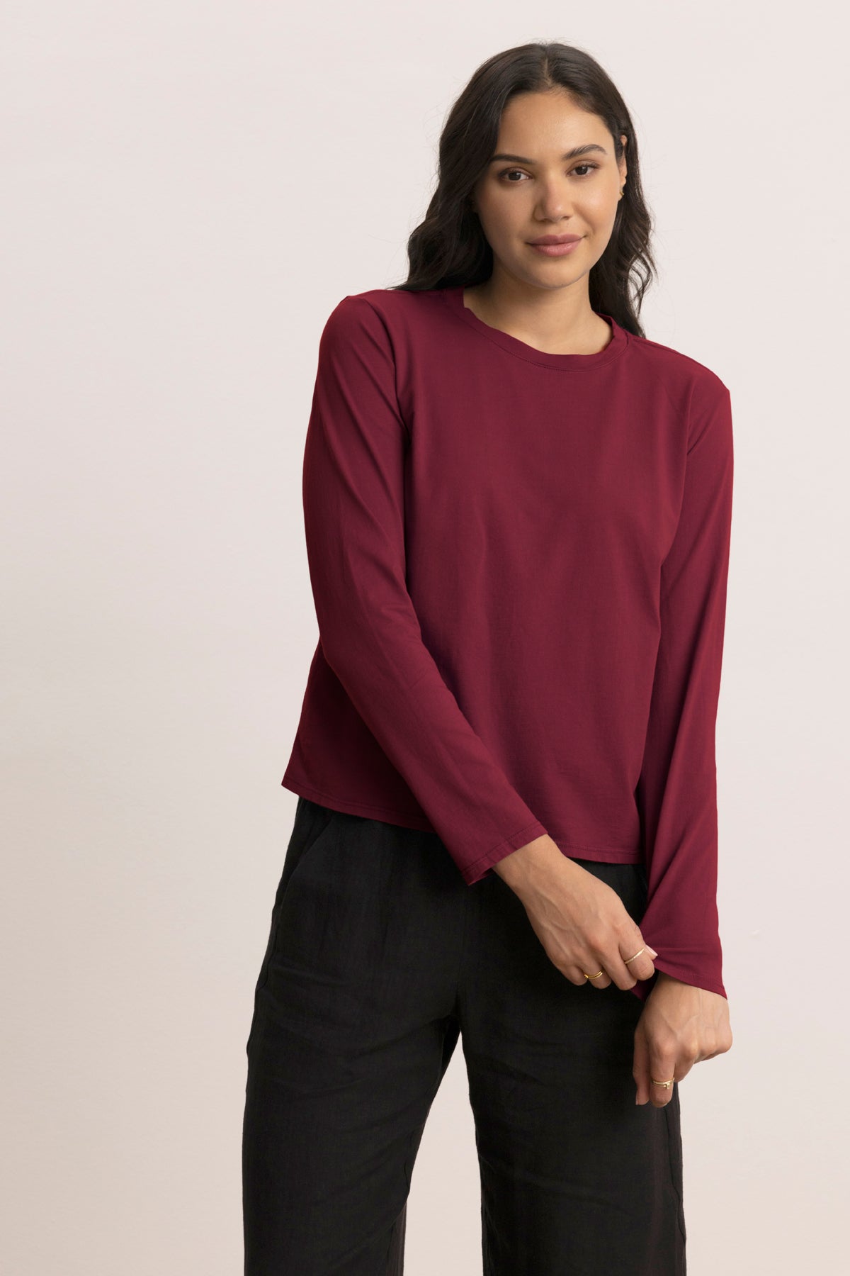 A woman with long dark hair wears the Velvet by Jenny Graham VICENTE TEE, a relaxed fit, long sleeve maroon top, paired with black pants, standing against a plain light background.-Hover Image