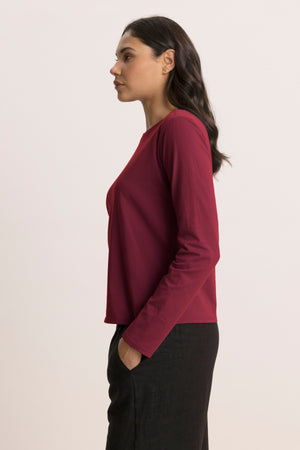 A woman stands in profile with her right hand in her pocket, wearing the Velvet by Jenny Graham VICENTE TEE—a relaxed fit long sleeve made from organic cotton—and black pants against a plain background.