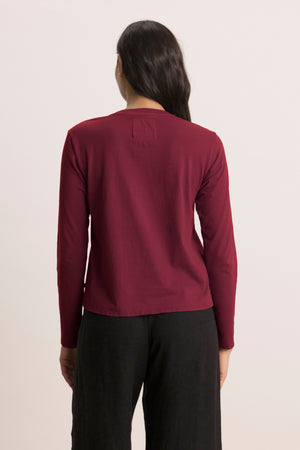 A person with long dark hair, seen from behind, wears the Velvet by Jenny Graham VICENTE TEE in maroon with black pants against a plain light background.