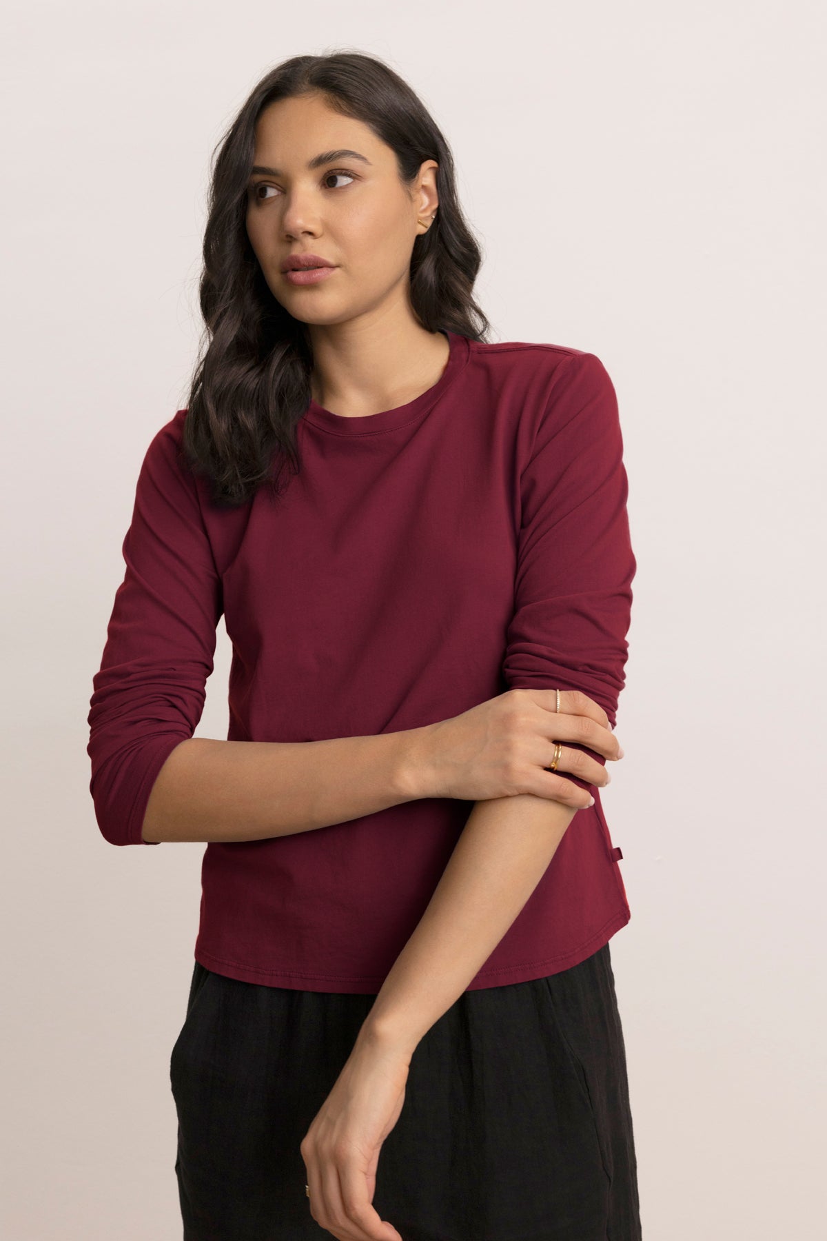 A woman with long dark hair wears the maroon VICENTE TEE by Velvet by Jenny Graham and black pants, standing against a light background and gazing slightly to the side.
