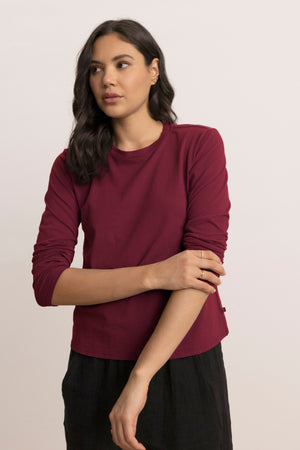 A woman with long dark hair wears the maroon VICENTE TEE by Velvet by Jenny Graham and black pants, standing against a light background and gazing slightly to the side.