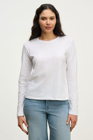 A woman with long dark hair wears the VICENTE TEE by Velvet by Jenny Graham—a relaxed fit, plain white long sleeve tee—paired with light blue jeans, standing against a neutral background.