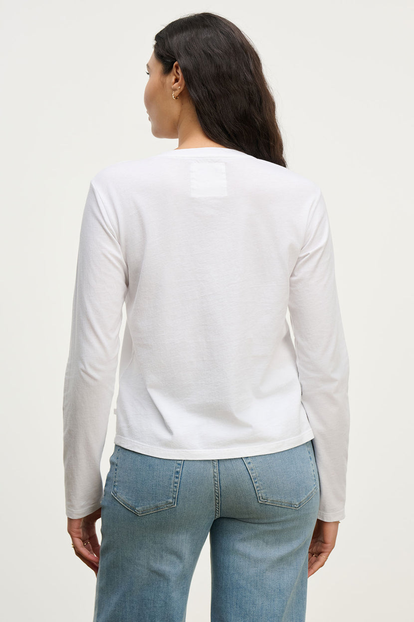 A woman with long dark hair is shown from the back wearing the Velvet by Jenny Graham VICENTE TEE, a relaxed fit organic cotton long sleeve shirt, paired with light blue jeans against a plain light background.