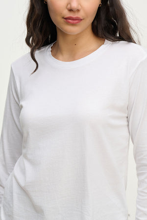 A woman wears the Velvet by Jenny Graham VICENTE TEE, a relaxed-fit long sleeve made from plain white organic cotton, shown from shoulders to waist with a neutral expression against a light background.