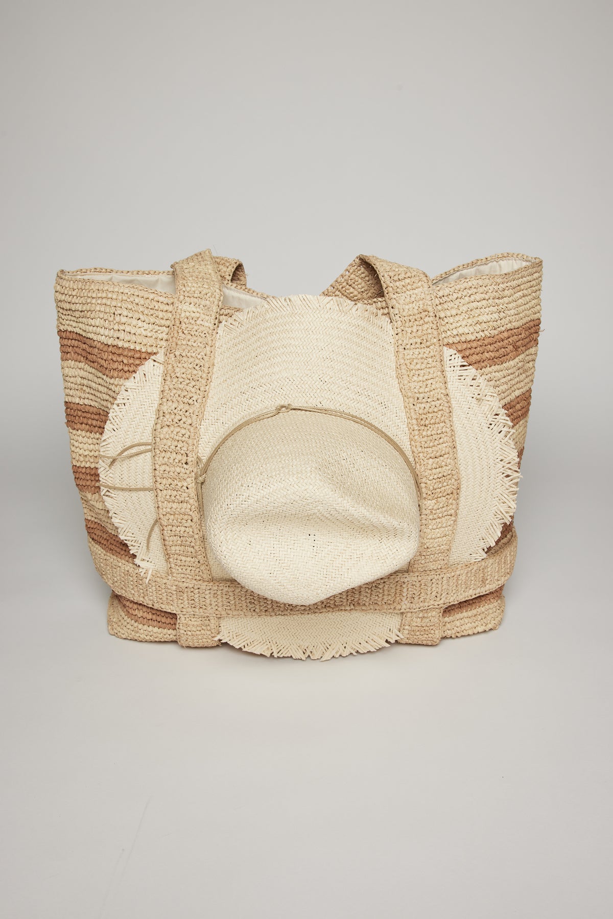   Woven raffia straw THE ORIGINAL STRAW TRAVELER BAG with wide beige straps and a pattern of horizontal tan and white stripes, displayed against a plain gray background by Velvet by Graham & Spencer. 