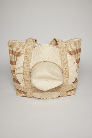 Woven raffia straw THE ORIGINAL STRAW TRAVELER BAG with wide beige straps and a pattern of horizontal tan and white stripes, displayed against a plain gray background by Velvet by Graham & Spencer.