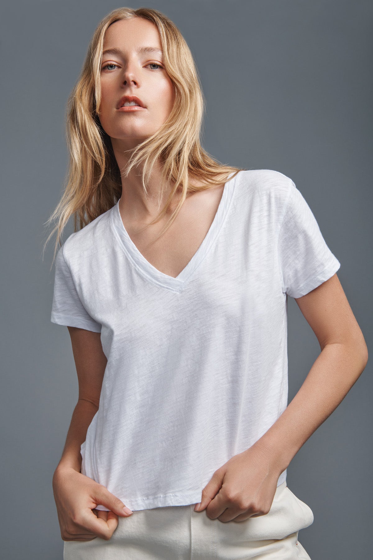 A woman with long blonde hair wears the ELISHA TEE by Velvet by Graham & Spencer—a plain white cotton slub V-neck tee—paired with light pants, standing against a gray background.-41776988324033