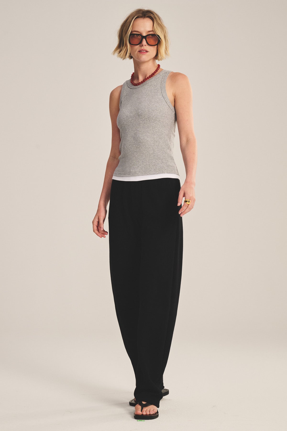 A person wears the ORION TANK TOP by Velvet by Graham & Spencer in gray, styled with black loose pants, black sandals, sunglasses, and a red necklace, standing against a plain background.