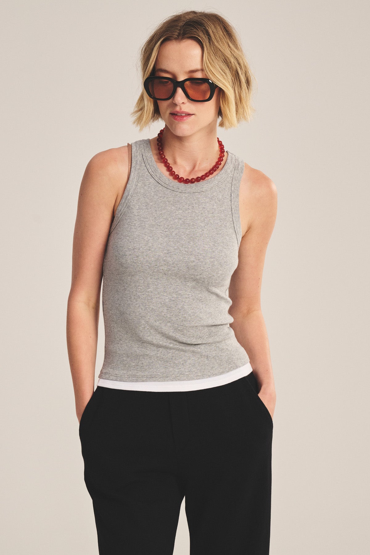 A woman stands with hands in her pockets, wearing Velvet by Graham & Spencer's ORION TANK TOP—a grey sleeveless style layered over a white ribbed tank—paired with black pants, sunglasses, and a red beaded necklace against a plain background.-41044068270273
