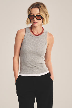 A woman stands with hands in her pockets, wearing Velvet by Graham & Spencer's ORION TANK TOP—a grey sleeveless style layered over a white ribbed tank—paired with black pants, sunglasses, and a red beaded necklace against a plain background.