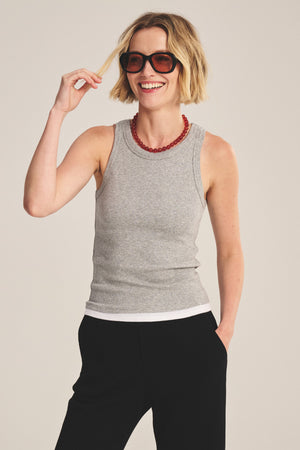 A woman with short blonde hair styles the ORION TANK TOP by Velvet by Graham & Spencer in grey over a white shirt, paired with black pants, dark sunglasses, and a red beaded necklace for a chic layered look. She smiles, hand raised, showing her fitted silhouette.