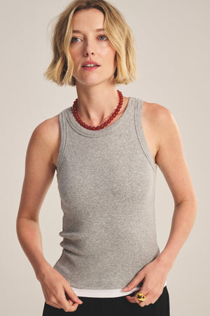 A person with short blonde hair wears the ORION TANK TOP by Velvet by Graham & Spencer, styled with black pants, a red beaded necklace, and a gold ring, standing against a neutral background.