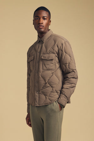 A man wearing the DANILO QUILTED JACKET by Velvet by Graham & Spencer—a light brown quilted shirt jacket that elevates men’s outerwear—paired with olive green pants stands against a beige background, looking at the camera with a neutral expression.