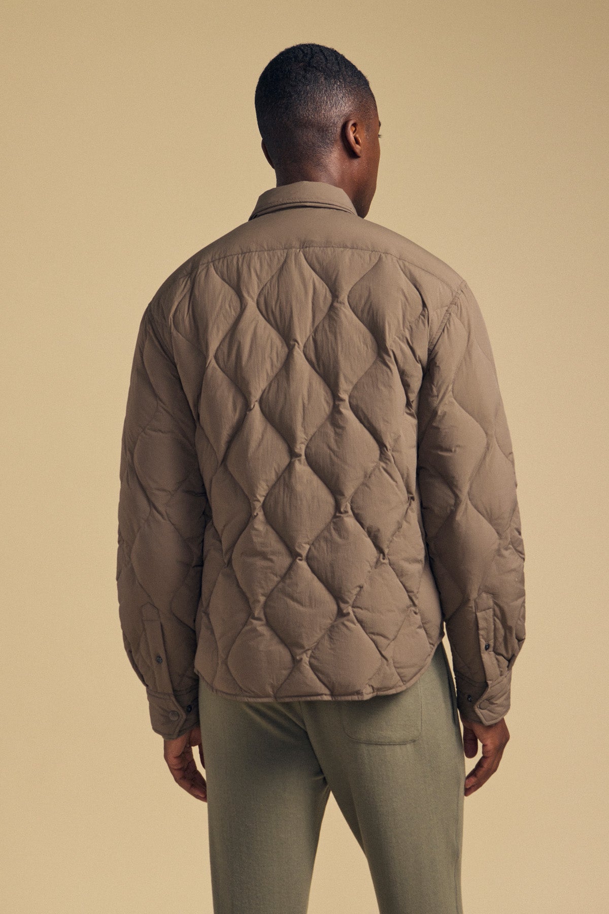A person stands facing away against a beige background, wearing the DANILO QUILTED JACKET by Velvet by Graham & Spencer in brown paired with light green pants, showcasing a stylish take on men’s outerwear.