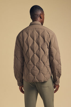 A person stands facing away against a beige background, wearing the DANILO QUILTED JACKET by Velvet by Graham & Spencer in brown paired with light green pants, showcasing a stylish take on men’s outerwear.