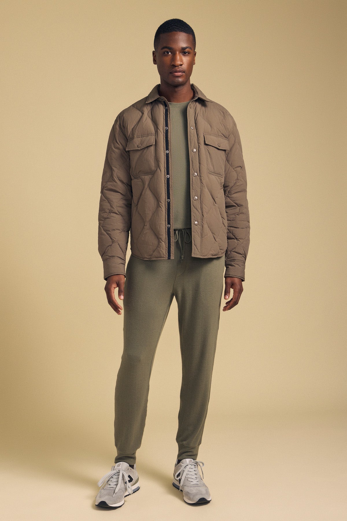 A man stands against a beige background, modeling the Velvet by Graham & Spencer DANILO QUILTED JACKET in brown nylon over an olive green sweatshirt and joggers, completed with gray sneakers.-42092460212417