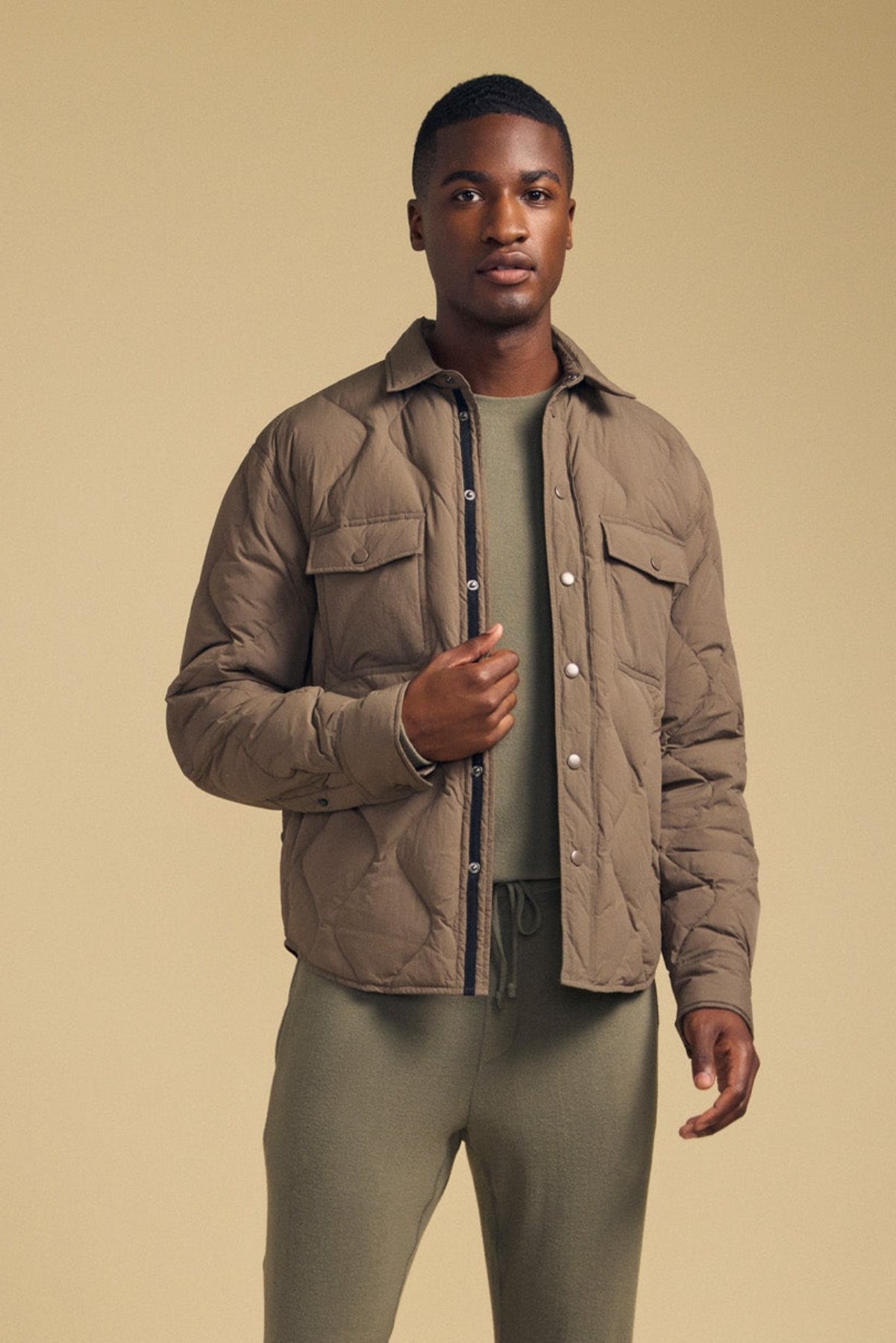 Wearing the DANILO QUILTED JACKET by Velvet by Graham & Spencer, a person stands against a beige background with the taupe jacket layered over an olive green shirt and pants, looking directly at the camera.