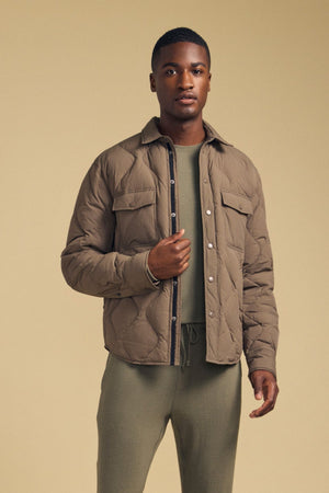 Wearing the DANILO QUILTED JACKET by Velvet by Graham & Spencer, a person stands against a beige background with the taupe jacket layered over an olive green shirt and pants, looking directly at the camera.