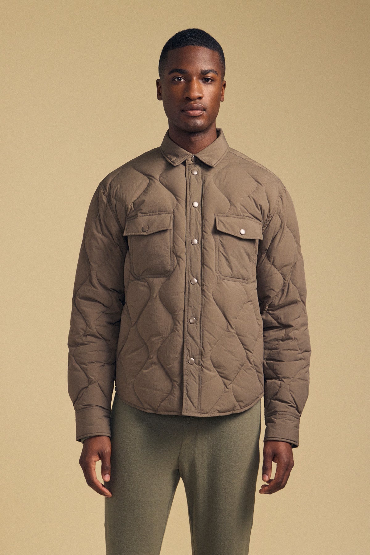 A man stands facing forward, wearing the DANILO QUILTED JACKET by Velvet by Graham & Spencer—a taupe shirt jacket with snap buttons and flap chest pockets—paired with light green pants, against a beige background.