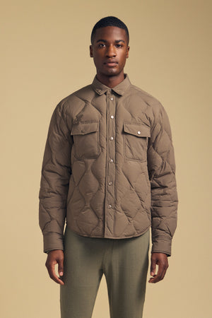 A man stands facing forward, wearing the DANILO QUILTED JACKET by Velvet by Graham & Spencer—a taupe shirt jacket with snap buttons and flap chest pockets—paired with light green pants, against a beige background.