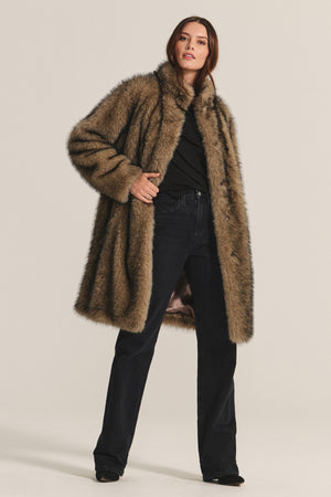 A person models the ADALYNN LUXE FAUX FUR JACKET by Velvet by Graham & Spencer—a long brown faux fur coat with a detachable faux leather belt—styled over a black top and dark jeans, perfect for chic cold-weather looks.