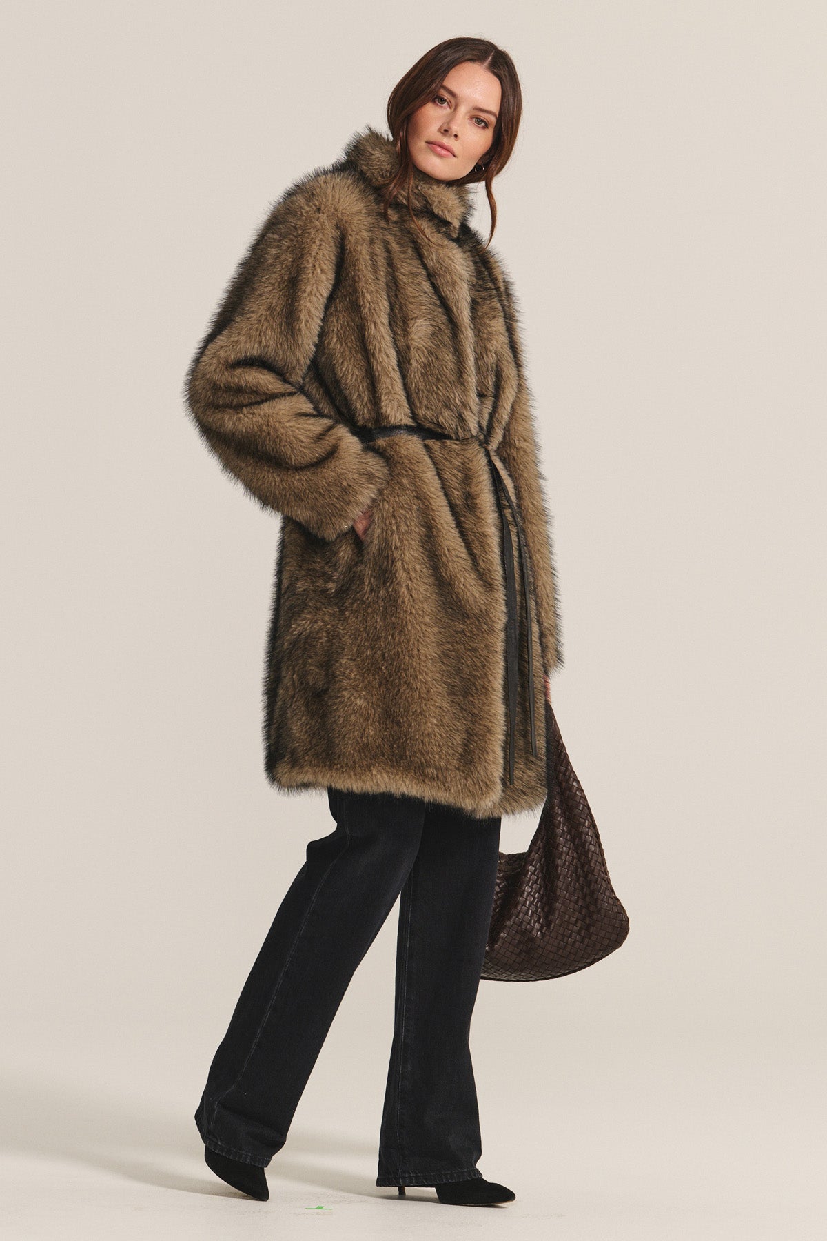 A woman wears the Velvet by Graham & Spencer ADALYNN LUXE FAUX FUR JACKET with a detachable faux leather belt, black pants, and black shoes, holding a large brown woven bag—all set against a plain light background.-40263641956545