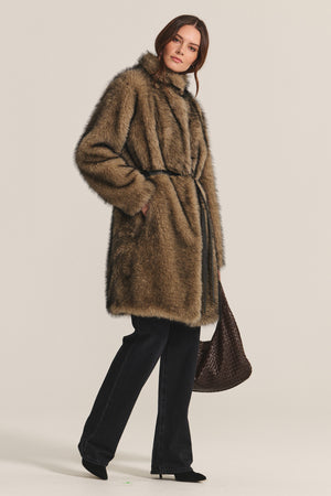 A woman wears the Velvet by Graham & Spencer ADALYNN LUXE FAUX FUR JACKET with a detachable faux leather belt, black pants, and black shoes, holding a large brown woven bag—all set against a plain light background.
