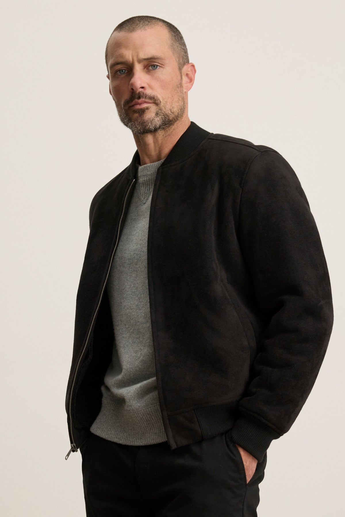 A man with short hair and a beard wears the AMIAS BOMBER JACKET by Velvet by Graham & Spencer over a gray sweater and black pants, standing against a plain light background.-40054203744449