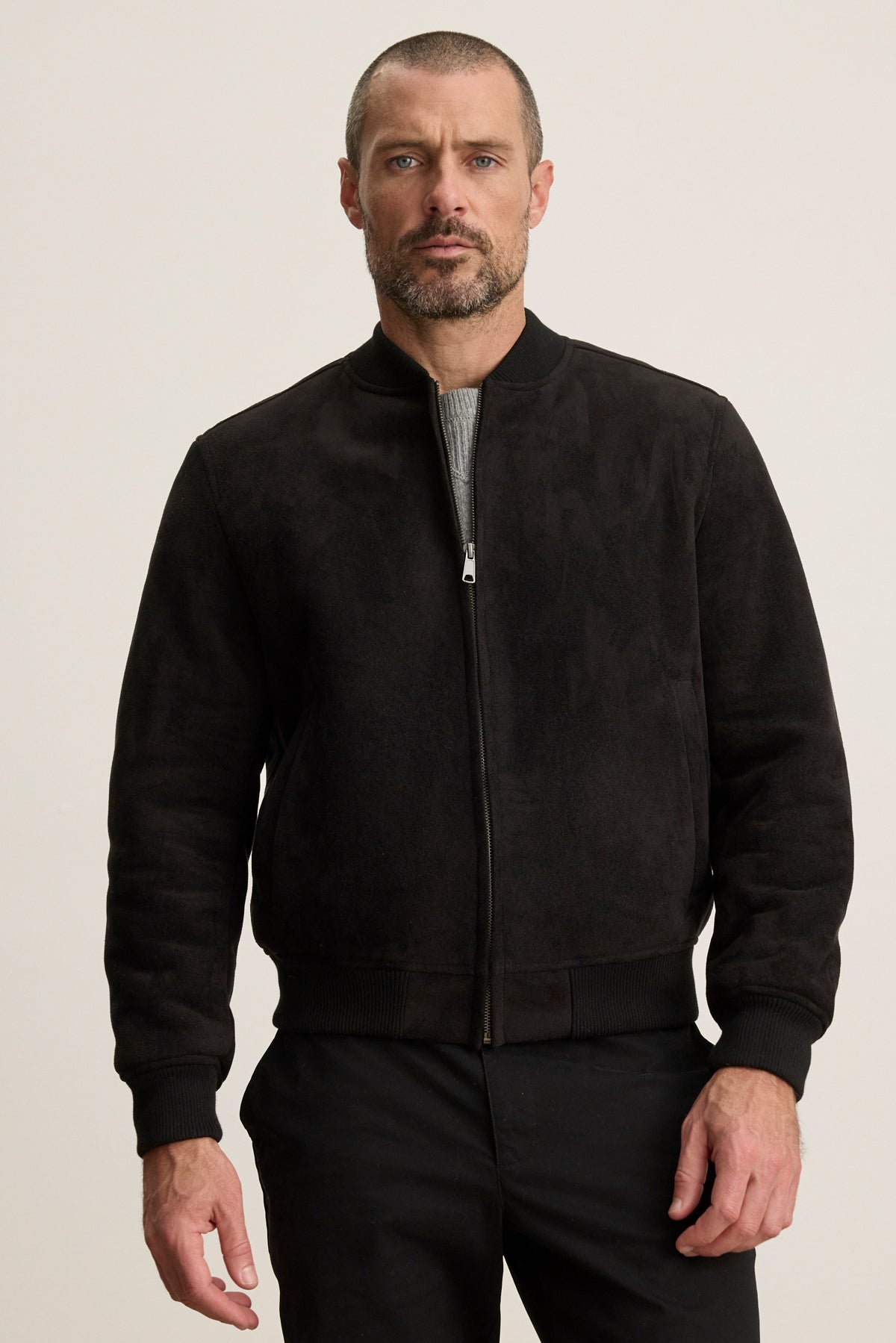 A man with a short beard and close-cropped hair models the AMIAS BOMBER JACKET by Velvet by Graham & Spencer in black faux suede over a gray shirt, set against a plain light background for a modern look.-40054203777217