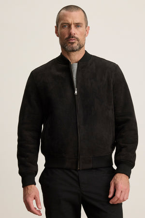 A man with a short beard and close-cropped hair models the AMIAS BOMBER JACKET by Velvet by Graham & Spencer in black faux suede over a gray shirt, set against a plain light background for a modern look.