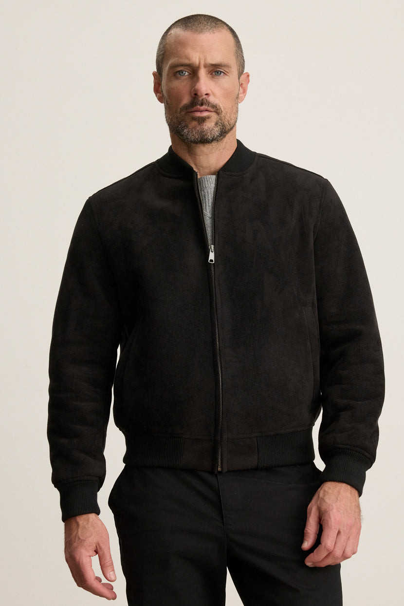 A man with a short beard and close-cropped hair models the AMIAS BOMBER JACKET by Velvet by Graham & Spencer in black faux suede over a gray shirt, set against a plain light background for a modern look.