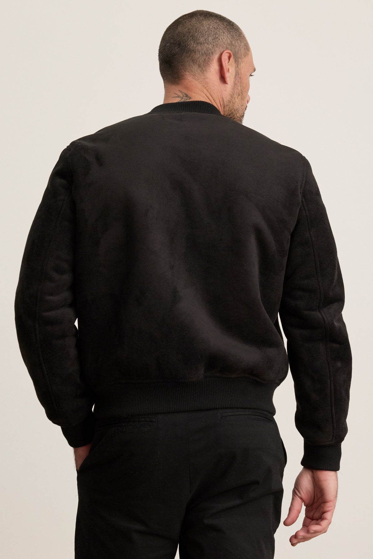 A man with short hair, seen from the back, models the AMIAS BOMBER JACKET by Velvet by Graham & Spencer and black pants against a light background, reflecting modern men's style.