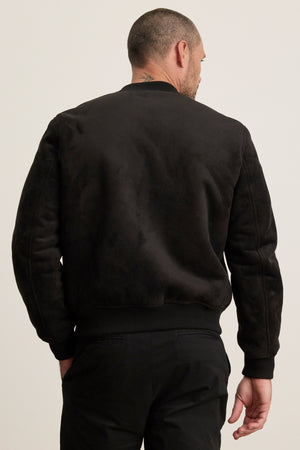A man with short hair, seen from the back, models the AMIAS BOMBER JACKET by Velvet by Graham & Spencer and black pants against a light background, reflecting modern men's style.