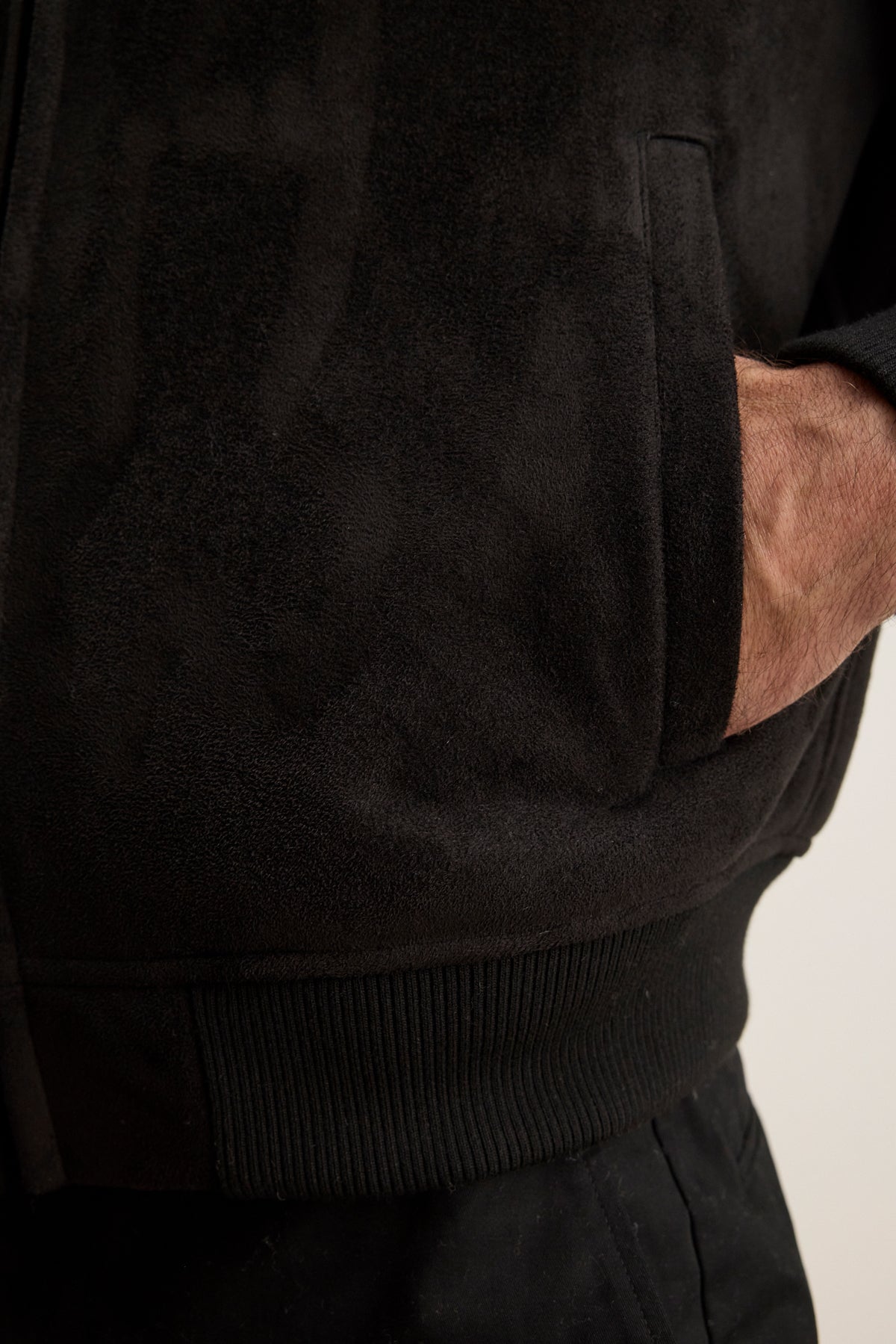 A person wearing the AMIAS BOMBER JACKET by Velvet by Graham & Spencer, a black faux suede piece, with their right hand in the front pocket, highlighting classic men's style.