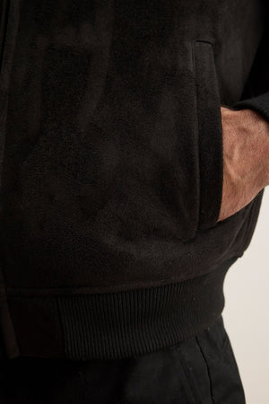 A person wearing the AMIAS BOMBER JACKET by Velvet by Graham & Spencer, a black faux suede piece, with their right hand in the front pocket, highlighting classic men's style.