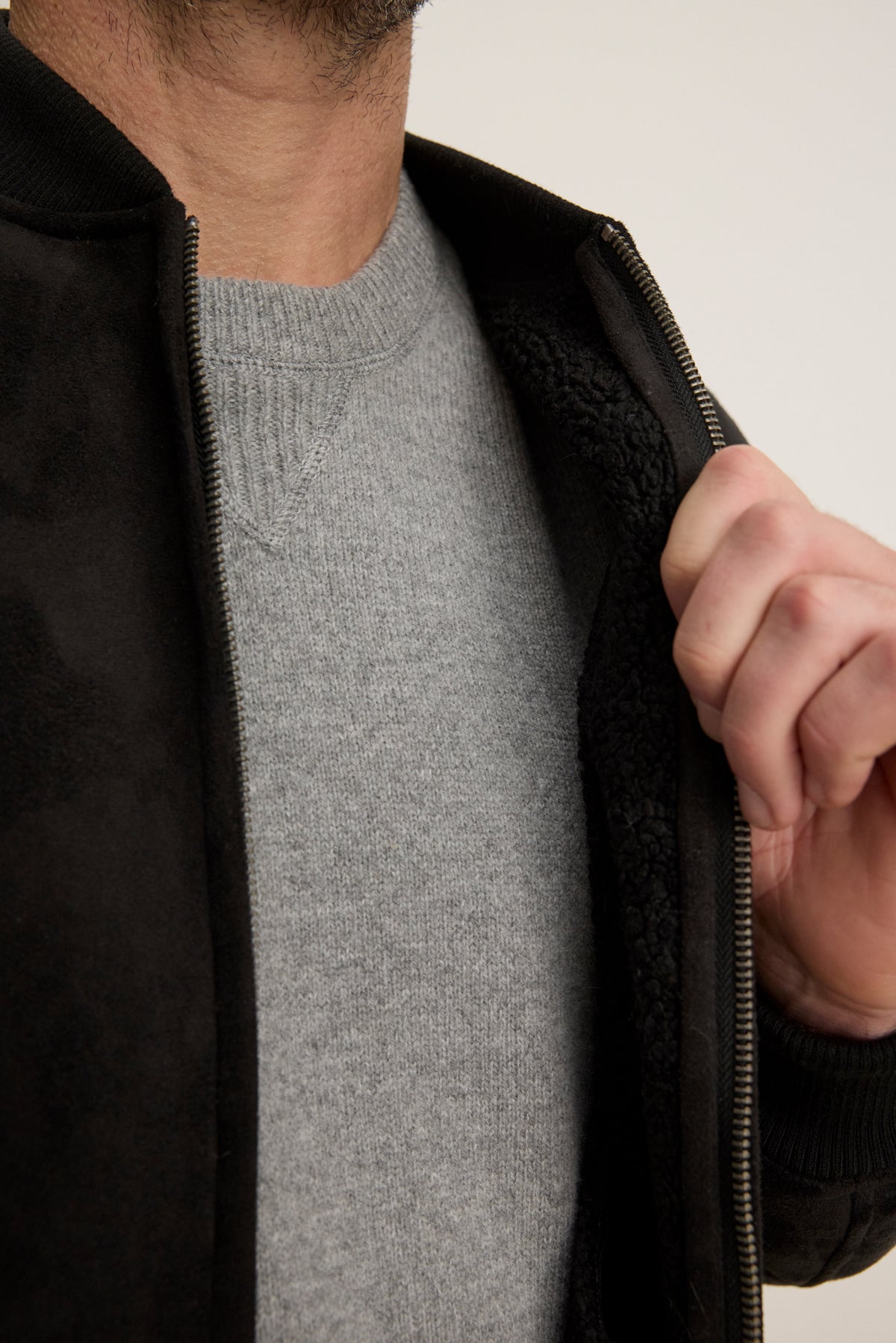 A person wearing a light grey sweater reveals the cozy sherpa lining of the partially unzipped AMIAS BOMBER JACKET by Velvet by Graham & Spencer.