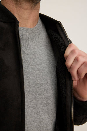 A person wearing a light grey sweater reveals the cozy sherpa lining of the partially unzipped AMIAS BOMBER JACKET by Velvet by Graham & Spencer.