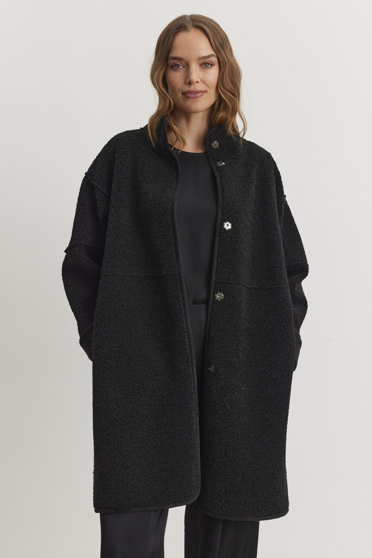 A person stands against a plain light background, wearing the CARA LUXE SHERPA REVERSIBLE JACKET from Velvet by Graham & Spencer—a luxurious reversible coat with sherpa detailing—over a sleek black outfit.-38219221893313