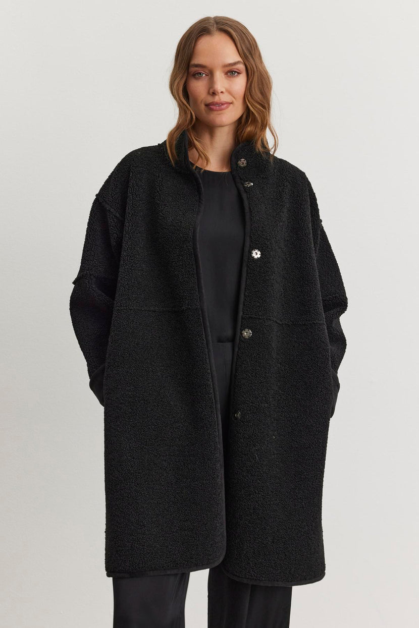 A person stands against a plain light background, wearing the CARA LUXE SHERPA REVERSIBLE JACKET from Velvet by Graham & Spencer—a luxurious reversible coat with sherpa detailing—over a sleek black outfit.