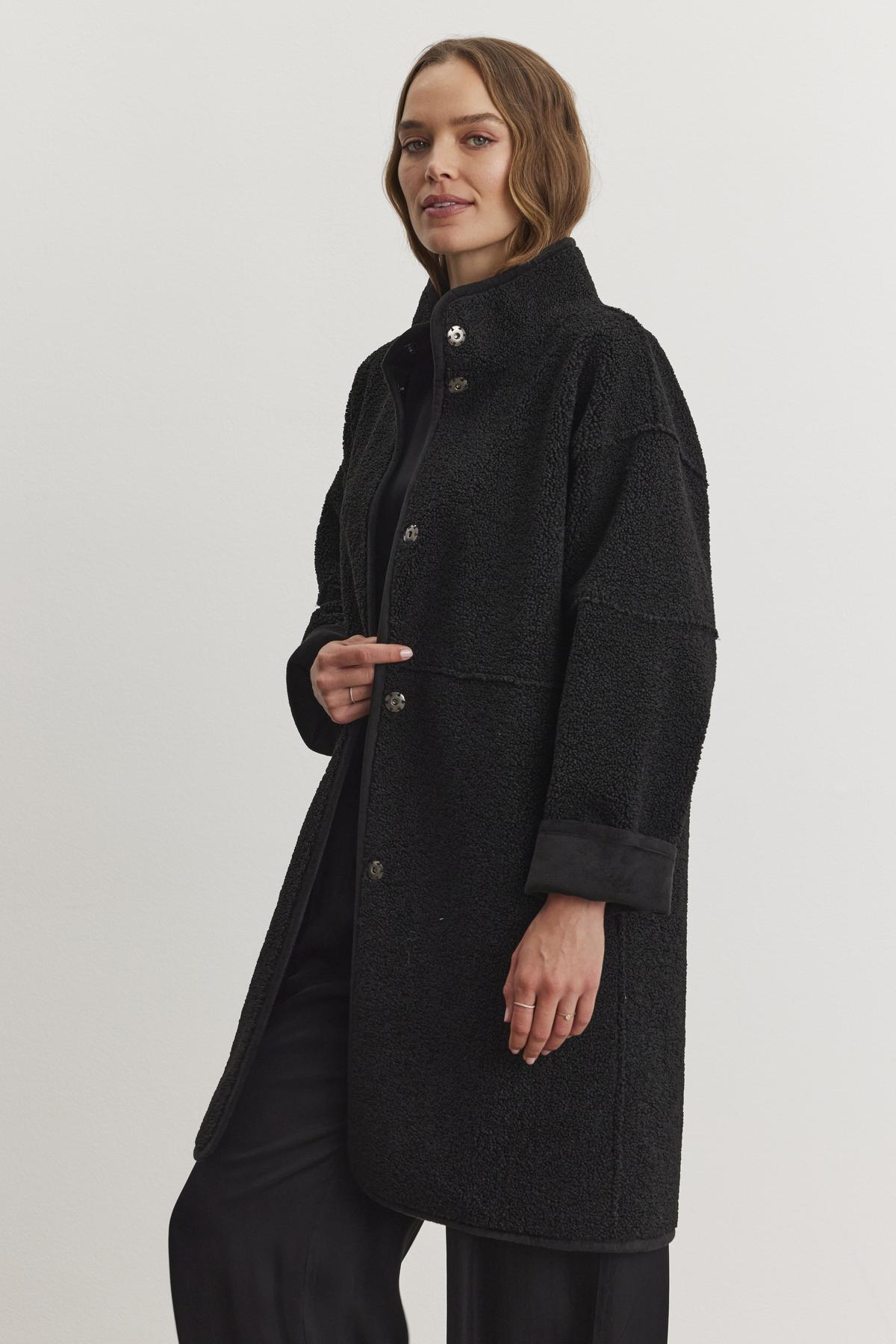   A person wearing a black Cara Luxe Sherpa Reversible Jacket by Velvet by Graham & Spencer, featuring a faux suede texture, stands against a plain background, looking forward. 