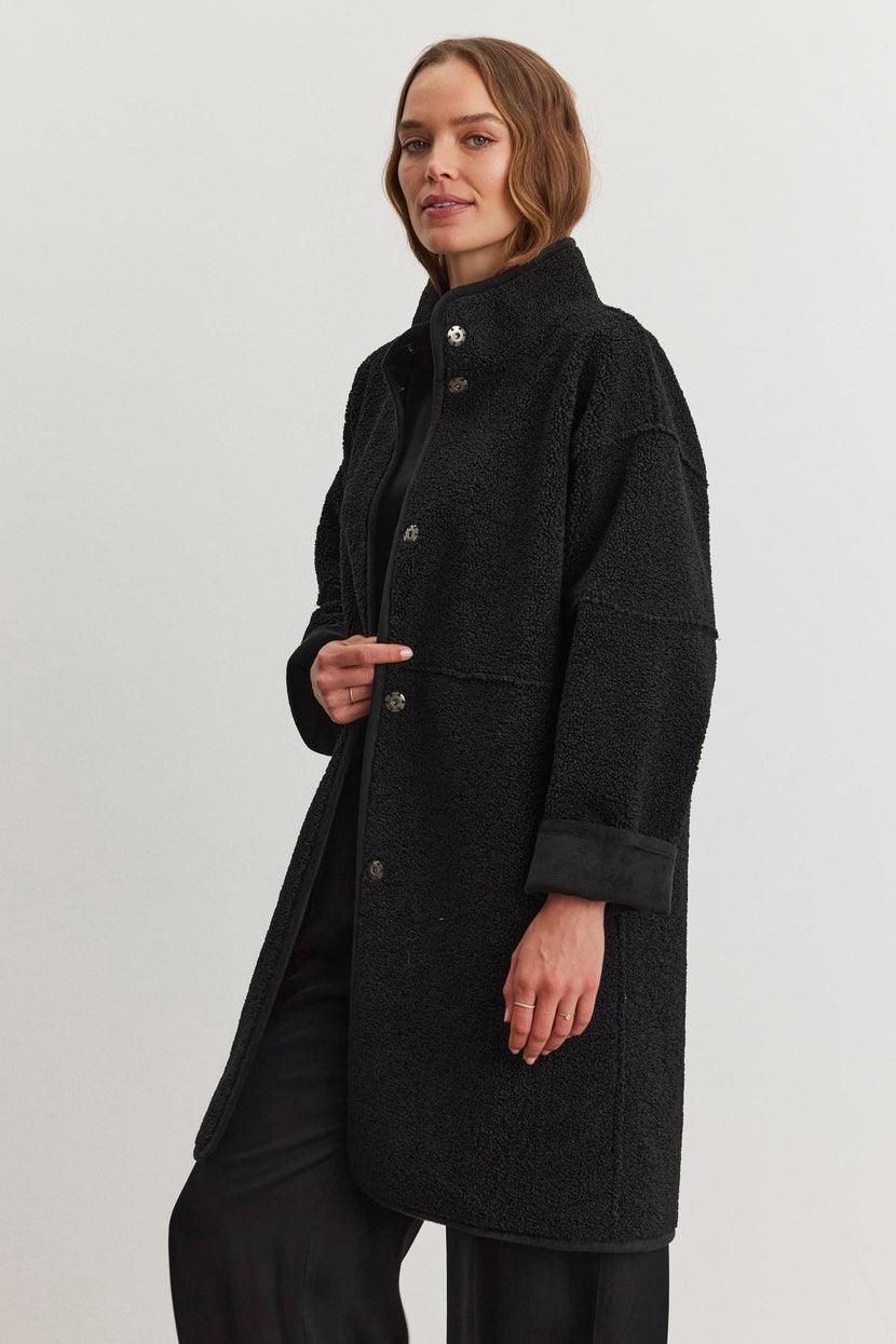 A person wearing a black Cara Luxe Sherpa Reversible Jacket by Velvet by Graham & Spencer, featuring a faux suede texture, stands against a plain background, looking forward.