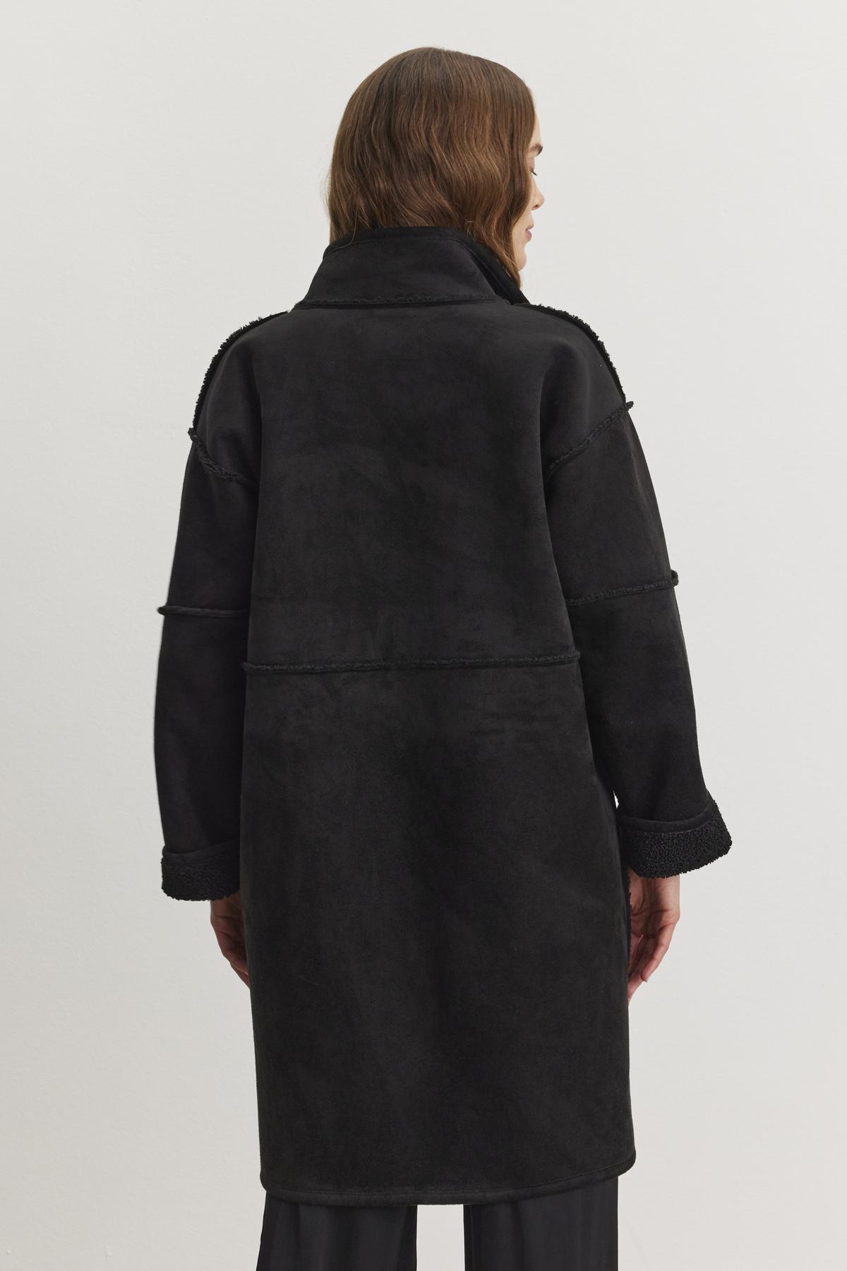   A person wearing a long black CARA LUXE SHERPA REVERSIBLE JACKET by Velvet by Graham & Spencer, shown from the back against a plain background, its subtle faux suede texture adding a touch of elegance. 