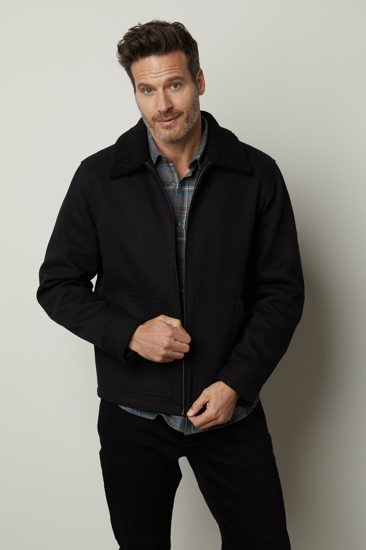 A man with short hair, wearing a black Velvet by Graham & Spencer CASH JACKET with a zipper front and a faux sherpa collar over a plaid shirt, stands in front of a neutral background, looking at the camera with a slight smile.-35472047046849