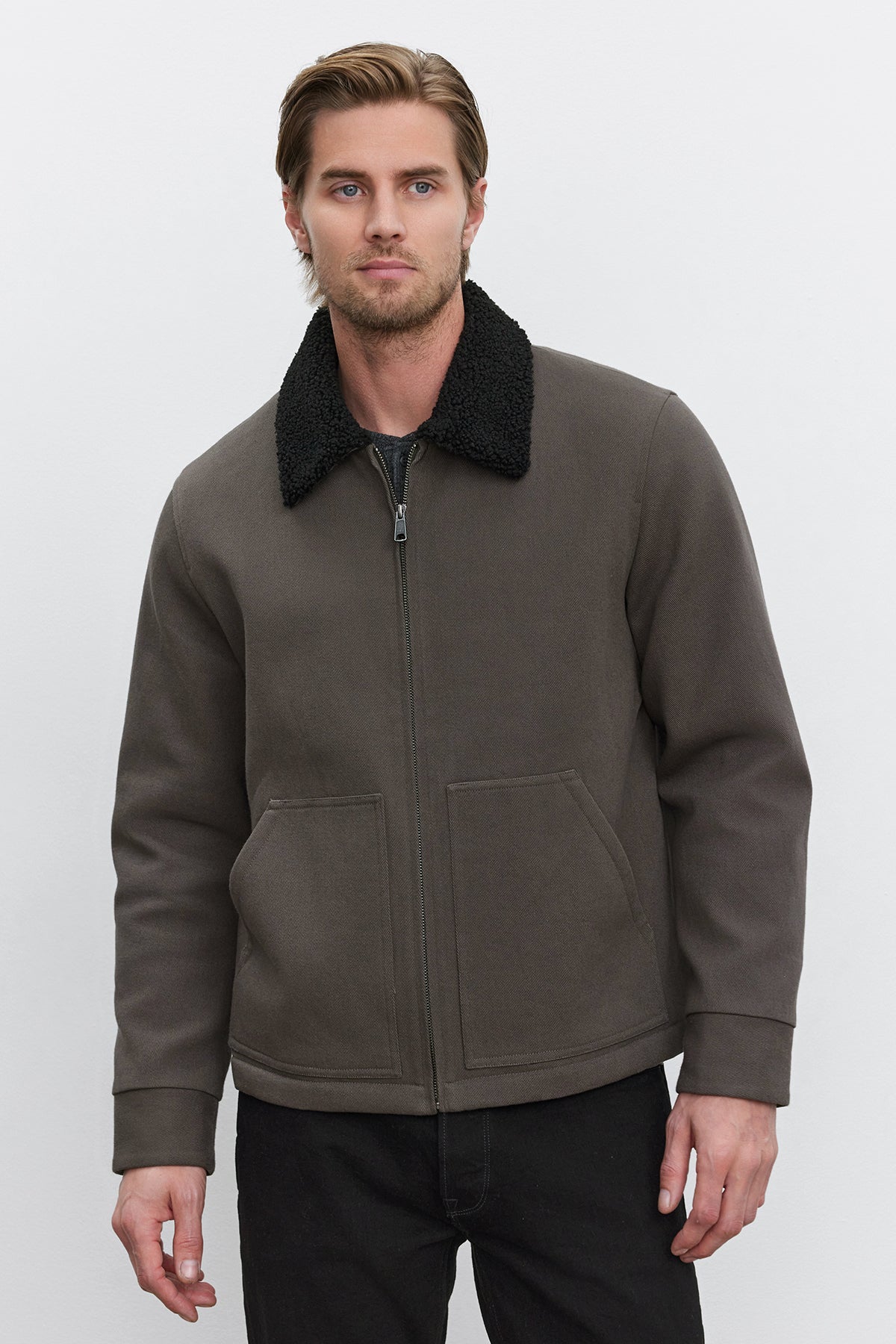   A man with short hair and a beard is wearing the CASH JACKET from Velvet by Graham & Spencer, which features a dark-colored, full-zip design with a black fleece collar. The durable cotton blend twill fabric enhances the rugged aesthetic as he stands facing forward against a plain white background. 