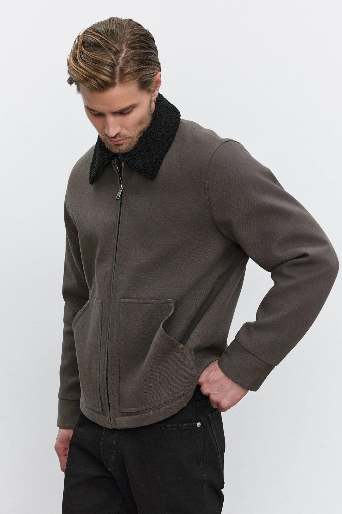   Man with light brown hair wearing the CASH JACKET by Velvet by Graham & Spencer, a dark green full-zip jacket featuring a black faux sherpa collar, paired with black pants. He is looking down with one hand in his jacket pocket. 